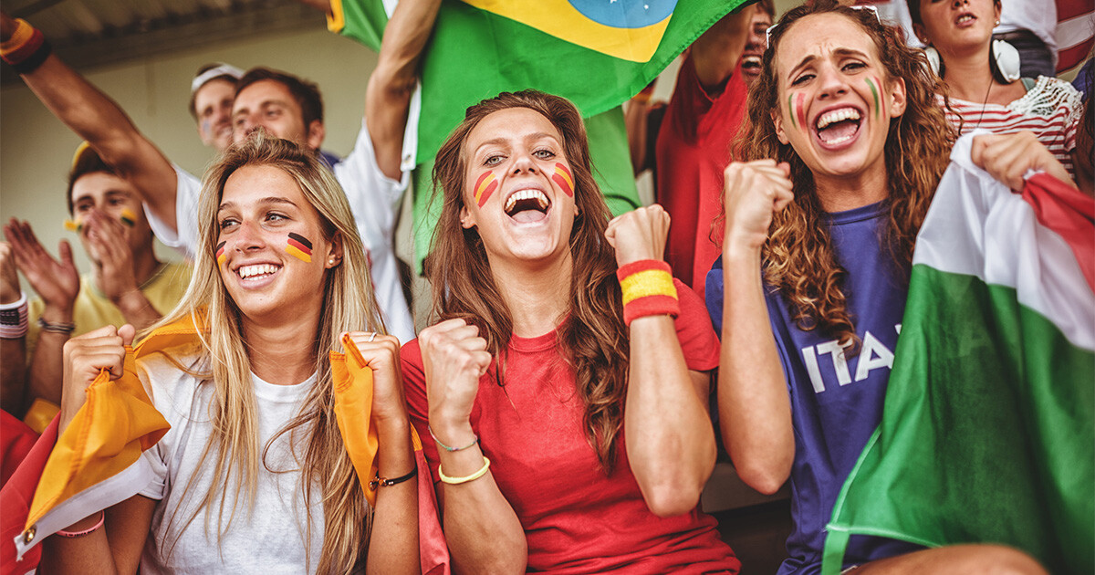 3 Frauen auf Zuschauertribüne unter anderen Fans. tragen deutsche, spanische und italienische Fankleidung