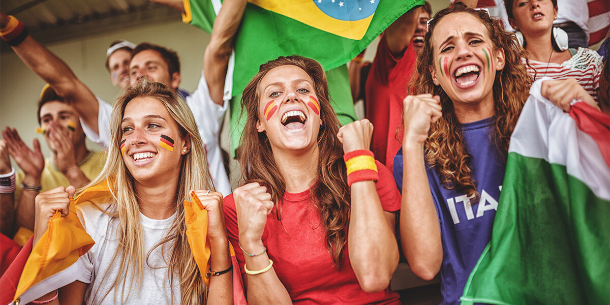 Drei Frauen auf Zuschauertribüne unter anderen Fans. tragen deutsche, spanische und italienische Fankleidung